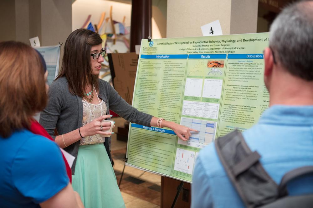 A student presenter holding a cup and pointing out something on her poster as she presents